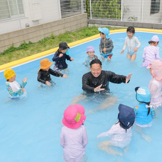 園内プールで先生を囲みながら水遊びをする園児たち