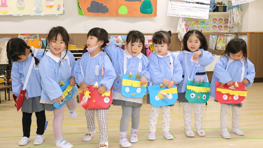 保育室で豆まき行事に参加し、手作りの鬼のバッグを身につけて並ぶ園児たち
